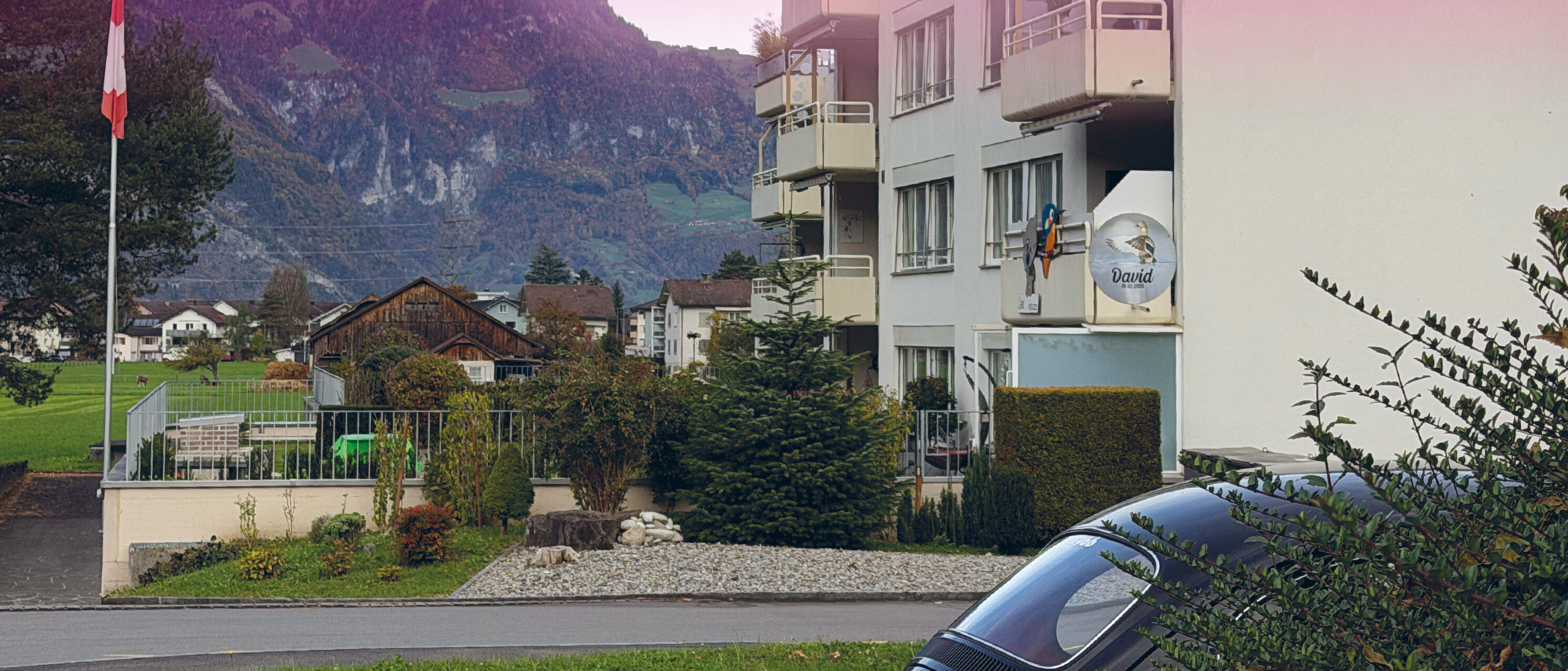Symbolbild: Ein Mehrfamilienhaus mit Balkonen in einem Wohnquartier. Vor dem Gebäude befinden sich Grünflächen und eine Strasse, im Hintergrund sind weitere Wohnhäuser und eine Berglandschaft zu sehen. Das Bild weist einen rosafarbenen Farbfilter auf.