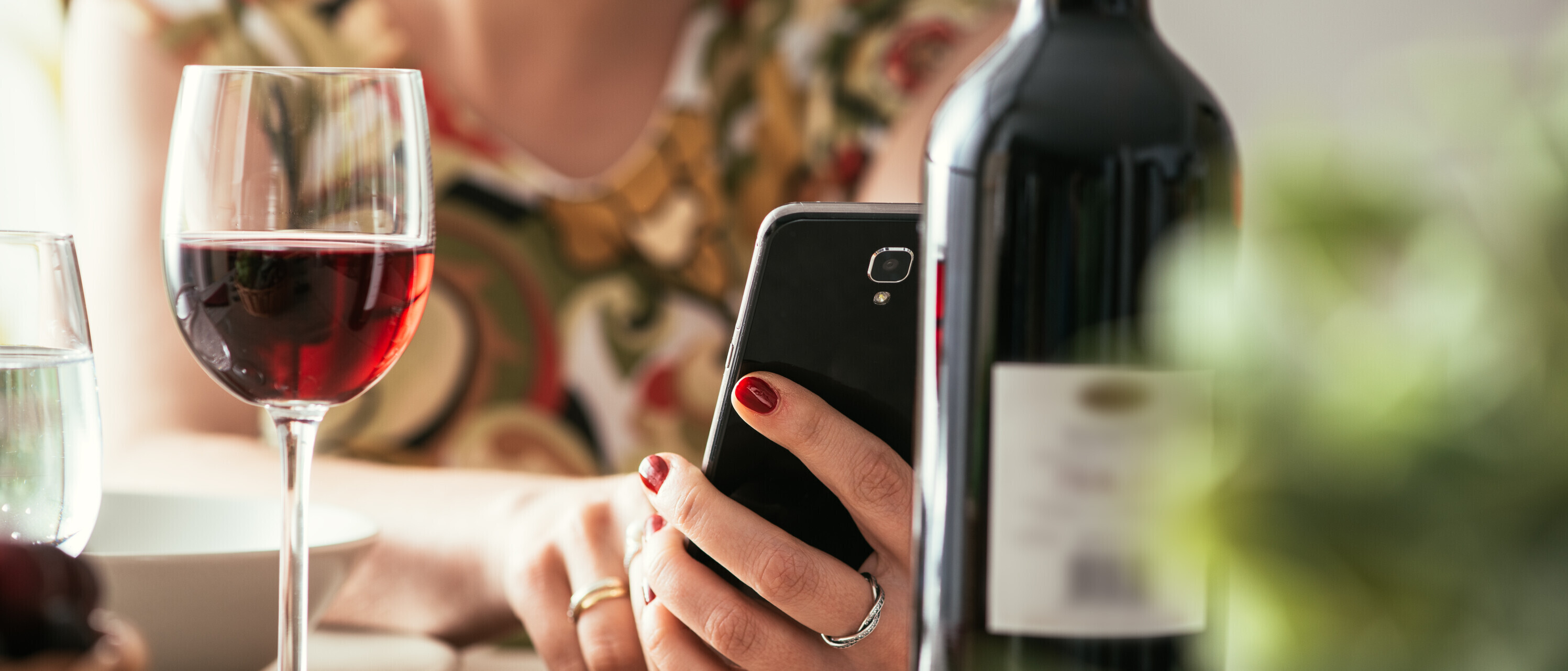Symbolbild: Eine Frau hält ihr Smartphone in der Hand und trink ein Glas Wein.