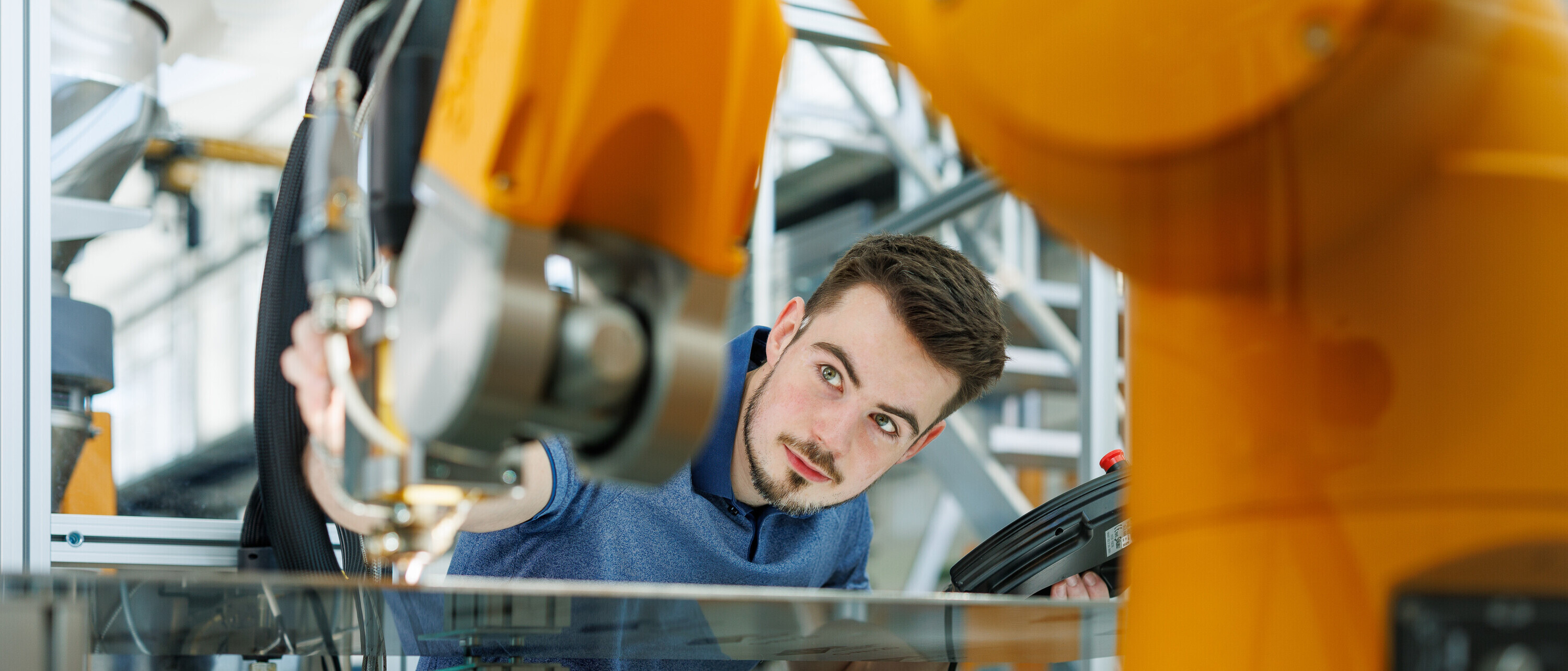 Junger Mann schaut zwischen Industrieroboter hindurch (Symbolbild für MSE Ingenieurstudium)
