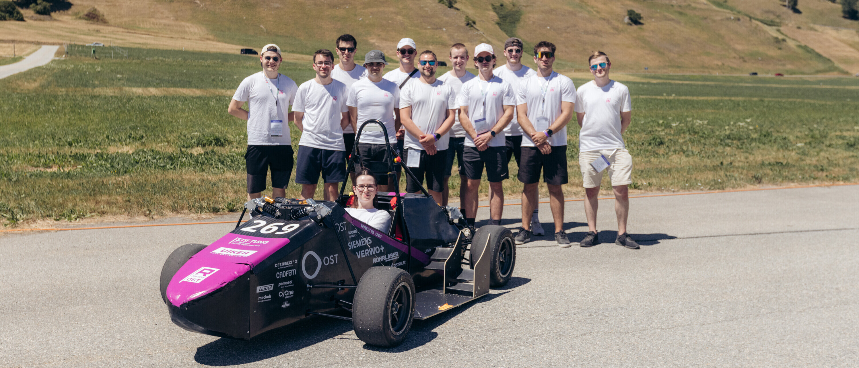 Eine Gruppe von Studierenden steht hinter einem schwarz-violetten Formula-Student-Rennwagen auf einer asphaltierten Strecke.