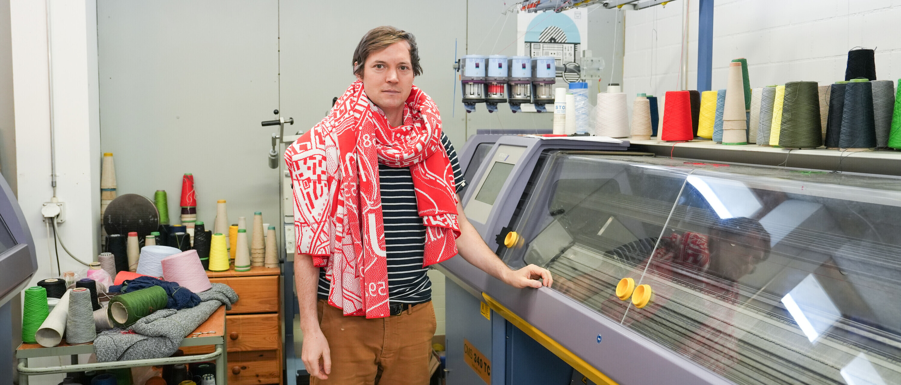 Urs Landis steht in seiner Strickerei neben einer Strickmaschine mit Fäden in verschiedenen Farben. Um seinen Hals trägt er einen rot-weissen Schal gewickelt.