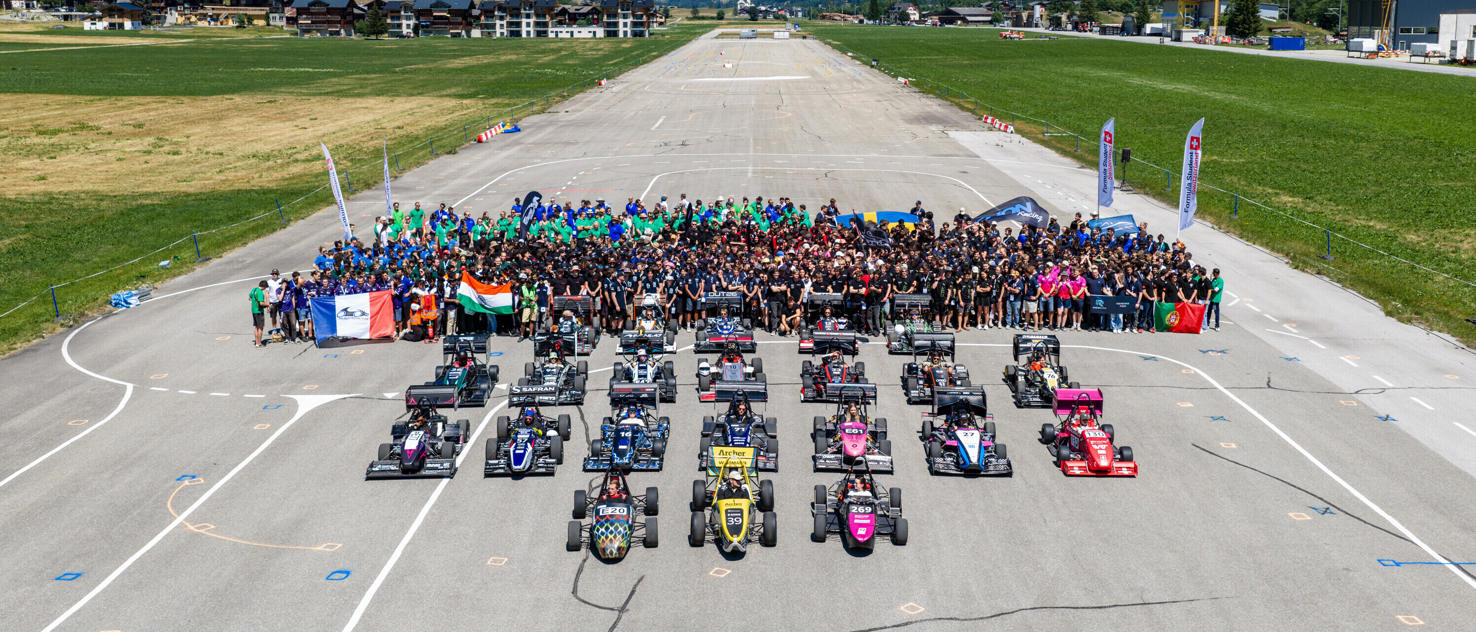 Gruppenbild der teilnehmenden Teams an der Formula Student Switzerland. Vor den Studierenden stehen zahlreiche Rennwagen auf einer Start- und Landebahn inmitten einer Berglandschaft.