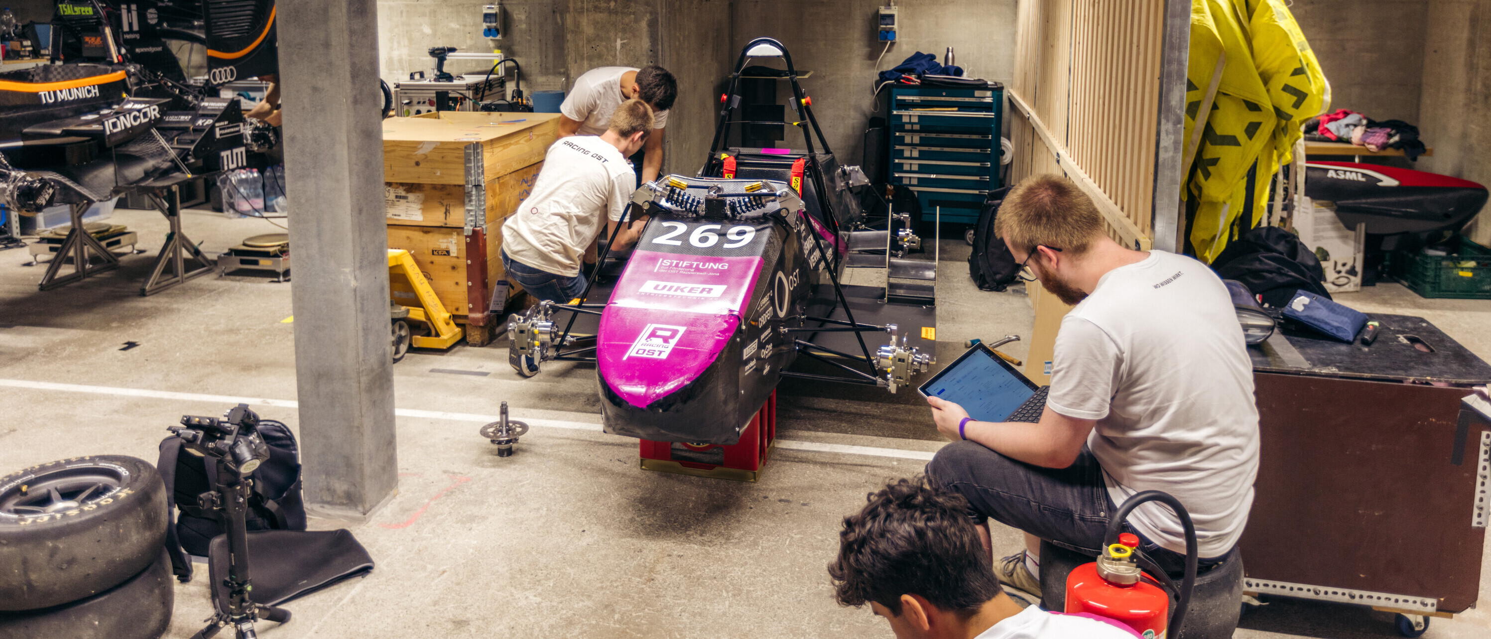 Mehrere Studierende arbeiten in einer Werkstatt an einem schwarz-violetten Formula-Student-Rennwagen. Eine Person sitzt mit einem Laptop neben dem Fahrzeug, während andere technische Arbeiten durchführen.