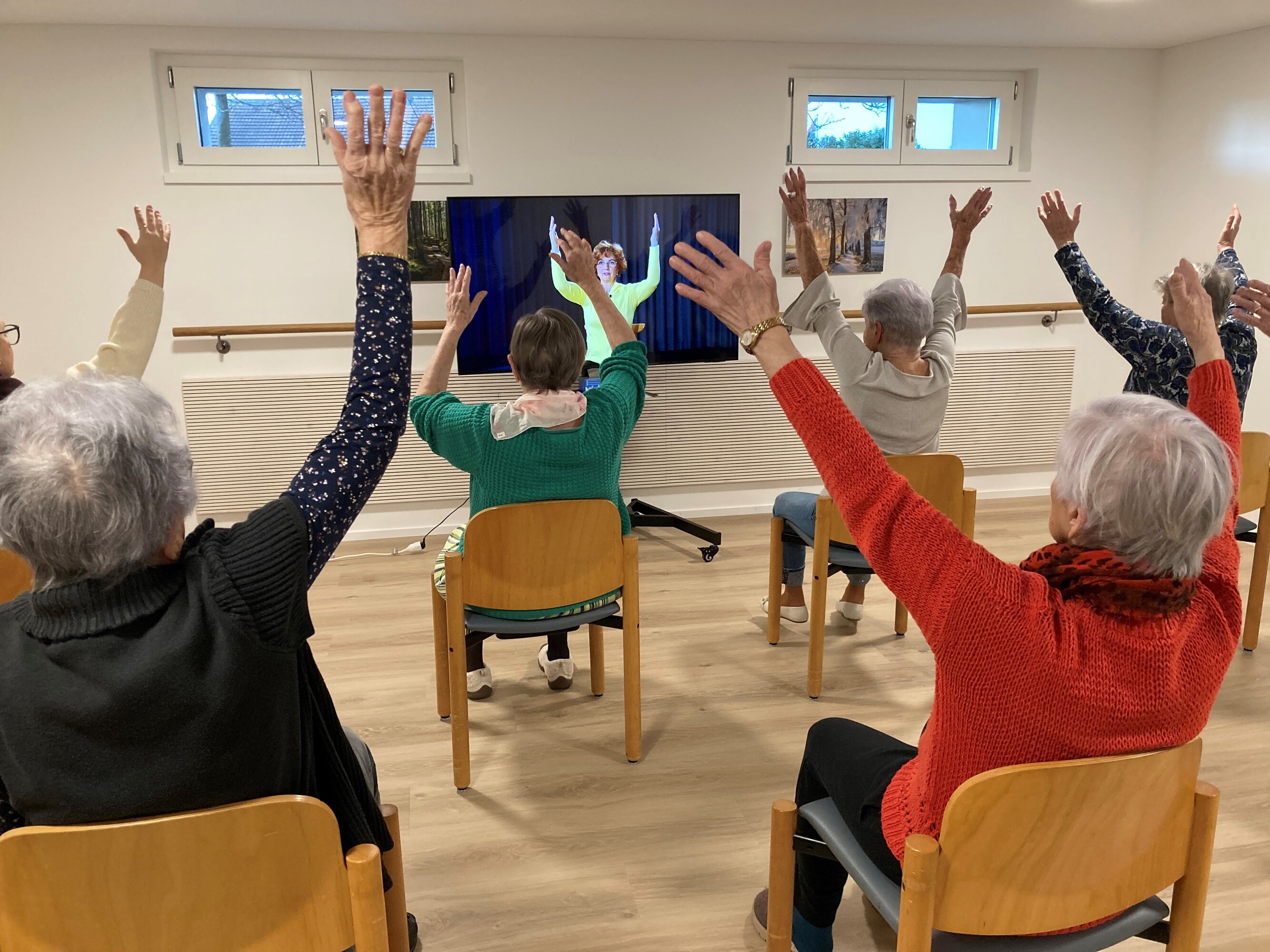 Fünf ältere Damen werden von hinten fotografiert, wie sie auf ihren Stühlen die Bewegungen aus dem Fernseher nachturnen.